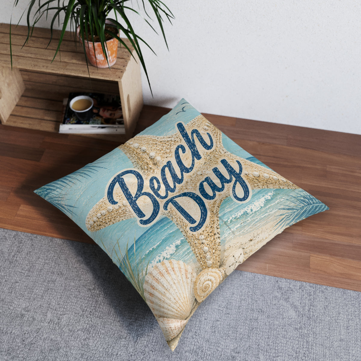 A square decorative throw pillow with a beach-themed print featuring a starfish, seashells, and the words 'Beach Day' in navy script, styled on a gray rug next to a wooden shelf with a plant.