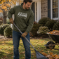 A man in an olive green hoodie with the text 'I'D Rather Be FISHING' rakes fallen leaves in a front yard next to a wheelbarrow of leaves.
