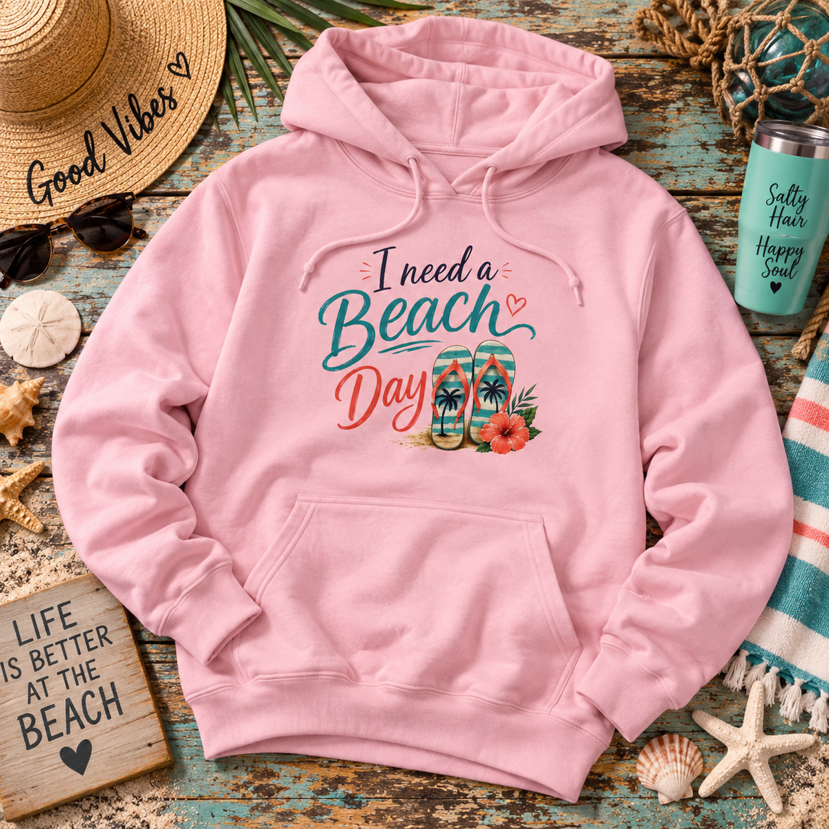 A pink pullover hoodie laid flat on a rustic blue wooden surface with beach-themed props including a straw hat, sunglasses, flip-flops, a tumbler, starfish, shells, and a sign that reads 'Life is Better at the Beach.' The hoodie has a large front graphic reading 'I need a Beach Day' with teal and coral lettering and a tropical flip-flop illustration.