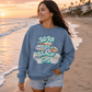 A woman stands on a sandy beach at sunset wearing a dusty blue crewneck sweatshirt with a colorful graphic that reads 'BORN TO BE A BEACH GIRL' and denim shorts.