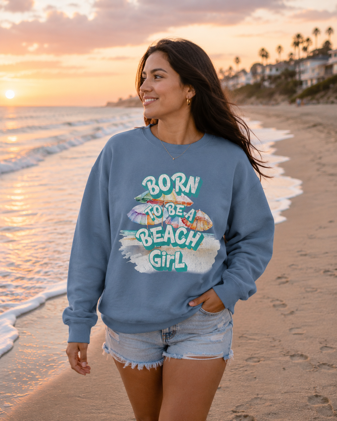 A woman stands on a sandy beach at sunset wearing a dusty blue crewneck sweatshirt with a colorful graphic that reads 'BORN TO BE A BEACH GIRL' and denim shorts.