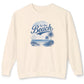 An off-white crewneck sweatshirt with a blue graphic print on the front that reads 'At the Beach' above a beach scene with palm trees and a sailboat.
