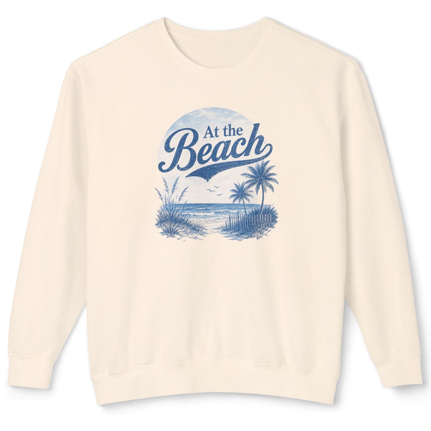 An off-white crewneck sweatshirt with a blue graphic print on the front that reads 'At the Beach' above a beach scene with palm trees and a sailboat.