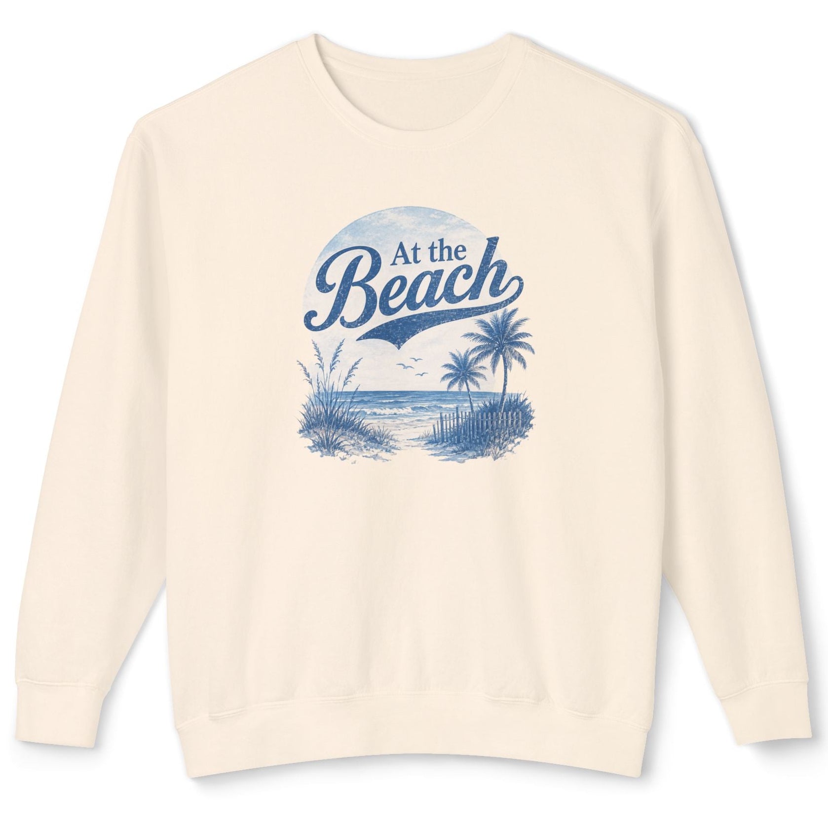 An off-white crewneck sweatshirt with a blue graphic print on the front that reads 'At the Beach' above a beach scene with palm trees and a sailboat.
