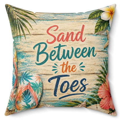 A square decorative throw pillow featuring a tropical beach design with palm leaves, flowers, and the text 'Sand Between the Toes' on a light wood-plank style background.