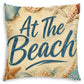 Square throw pillow featuring a beach-themed design with the phrase 'At The Beach' in dark blue script over a sandy beige background, decorated with starfish and seashells.