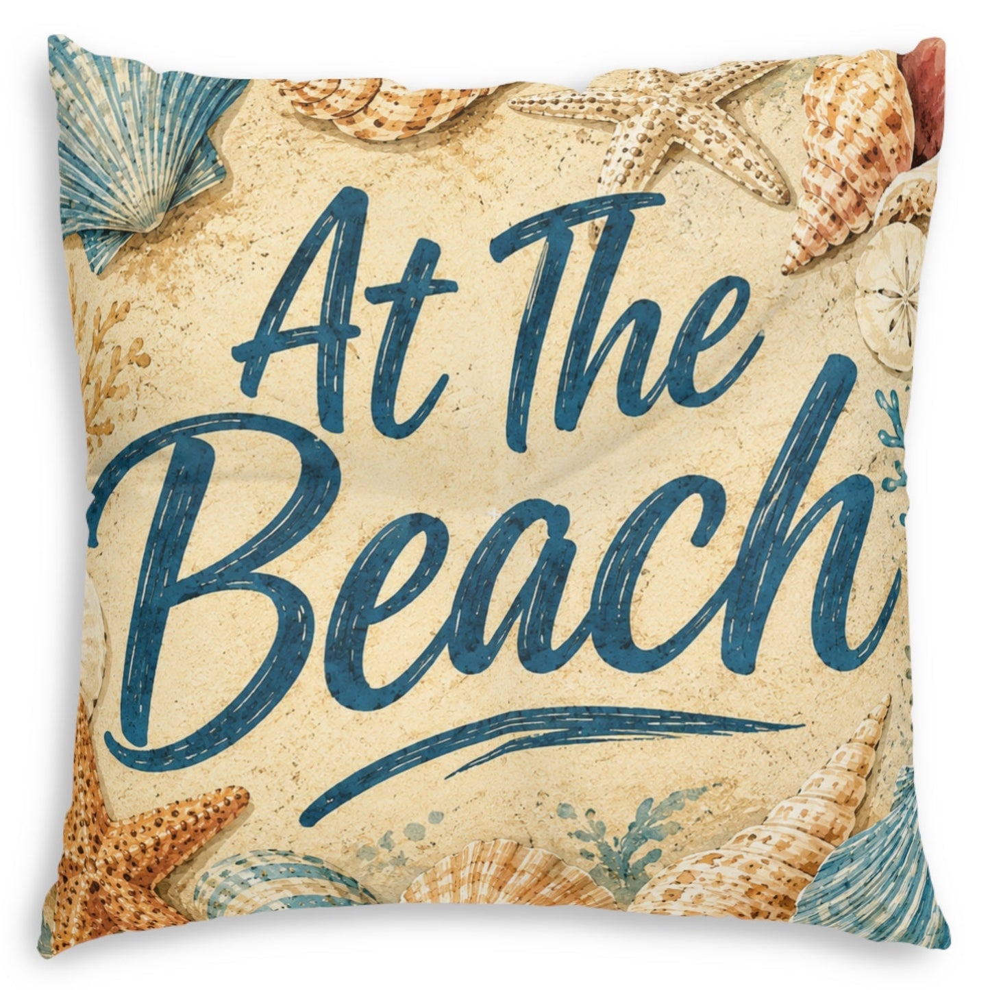 Square throw pillow featuring a beach-themed design with the phrase 'At The Beach' in dark blue script over a sandy beige background, decorated with starfish and seashells.