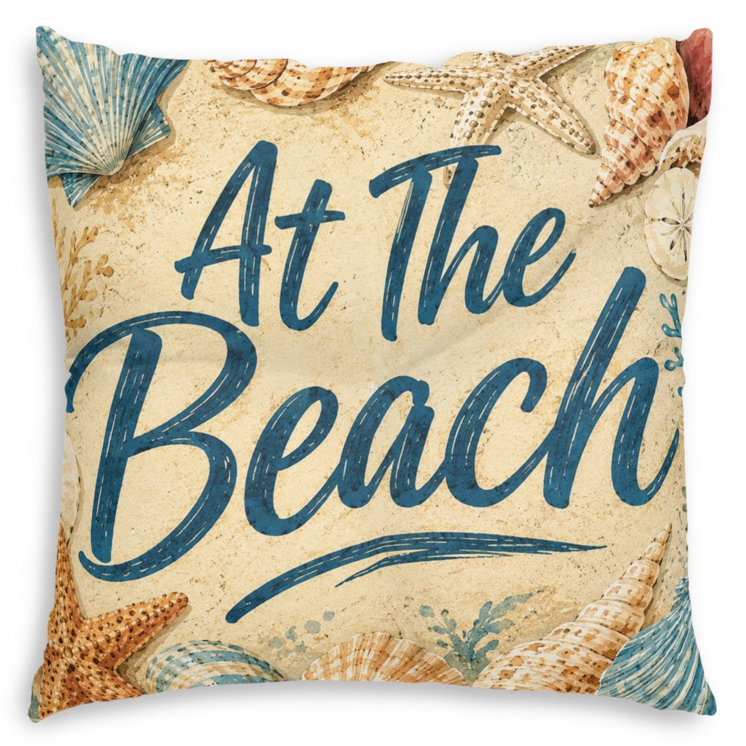 Square throw pillow featuring a beach-themed design with the phrase 'At The Beach' in dark blue script over a sandy beige background, decorated with starfish and seashells.
