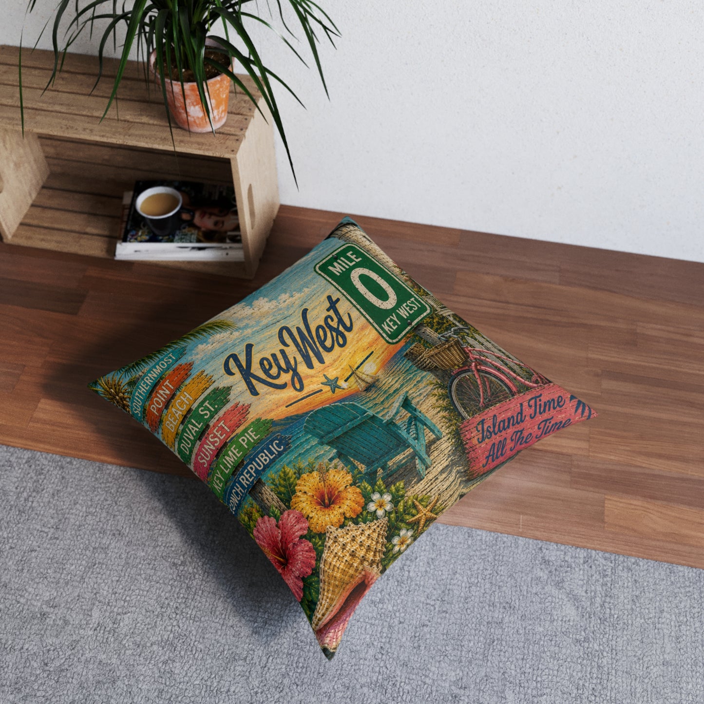 A square decorative throw pillow with a colorful vintage-style 'Key West' travel poster print, styled on a gray rug next to a wooden bench with a potted plant.