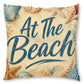 Square decorative throw pillow featuring the phrase 'At The Beach' in dark blue script over a sandy beige background, decorated with illustrated seashells and starfish.