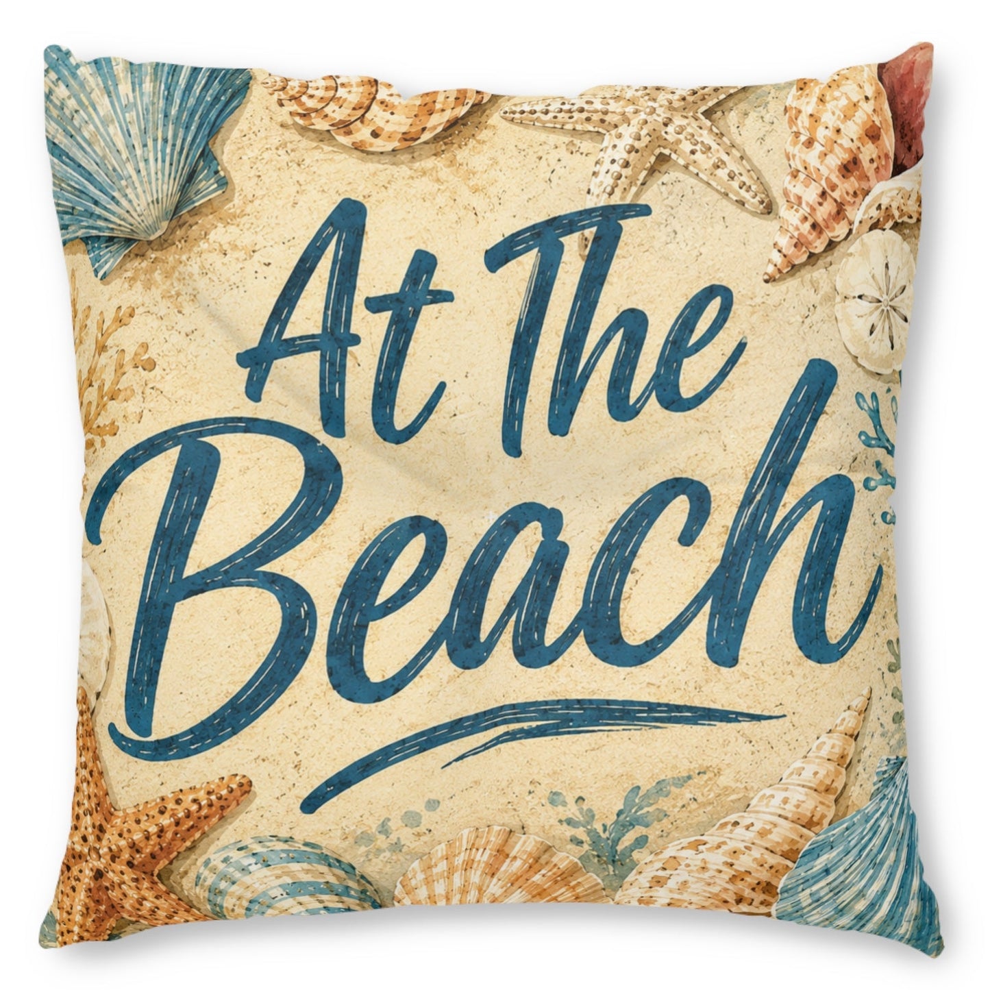 Square decorative throw pillow featuring the phrase 'At The Beach' in dark blue script over a sandy beige background, decorated with illustrated seashells and starfish.