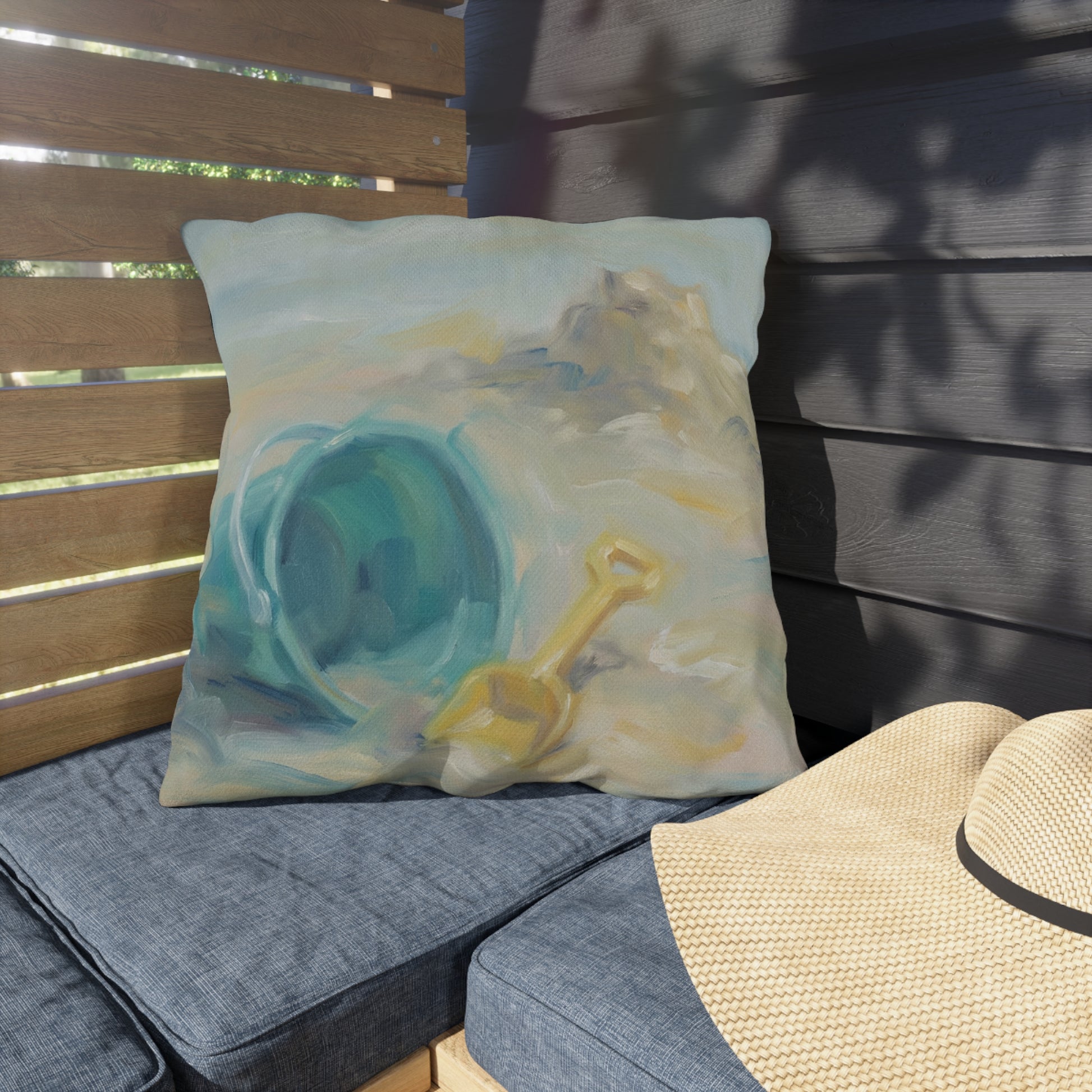 Decorative pillow with a beach scene featuring a blue bucket and yellow shovel on a wooden bench.