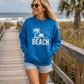A woman wearing a bright blue crewneck sweatshirt with a white palm tree and 'BEACH' graphic, walking on a wooden boardwalk at the beach.
