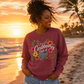 A woman stands on a beach at sunset wearing a dusty rose long-sleeve graphic sweatshirt with the text 'I'm in a Caribbean State of Mind' surrounded by tropical flowers, paired with light-wash denim shorts.