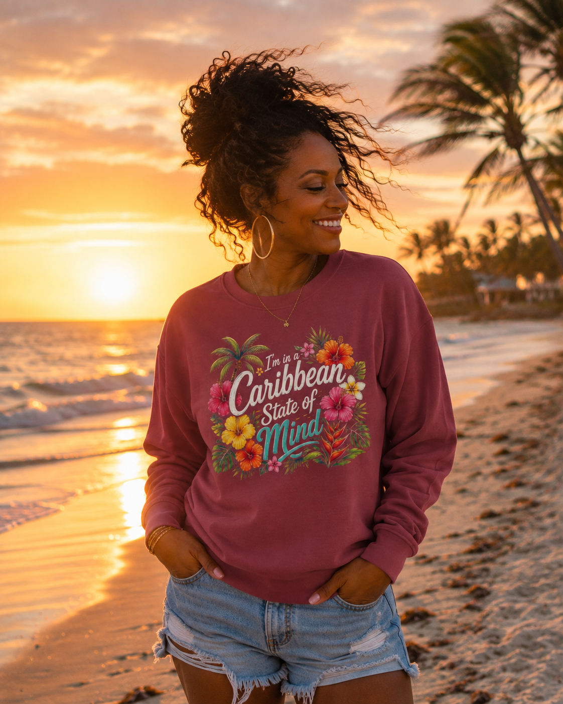 A woman stands on a beach at sunset wearing a dusty rose long-sleeve graphic sweatshirt with the text 'I'm in a Caribbean State of Mind' surrounded by tropical flowers, paired with light-wash denim shorts.