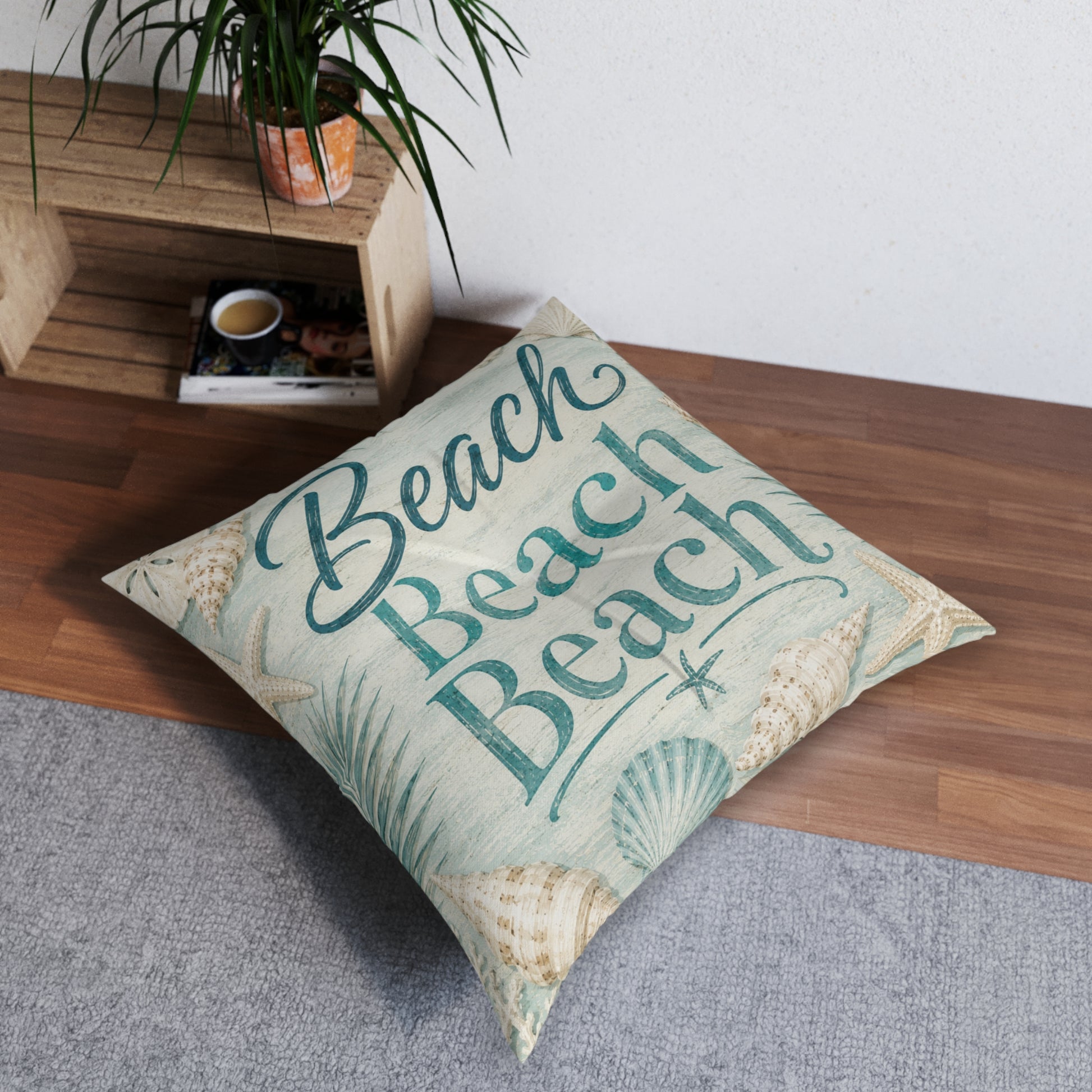 Square floor pillow with teal “Beach” text and seashell accents, angled on carpet and wood