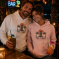 A man and a woman sitting together at a bar, both wearing matching light-colored hoodies with a tropical tiki graphic that reads 'TIKI VIBES'. The man wears a white hoodie and holds a tiki mug, while the woman wears a light pink hoodie and holds a cocktail glass.
