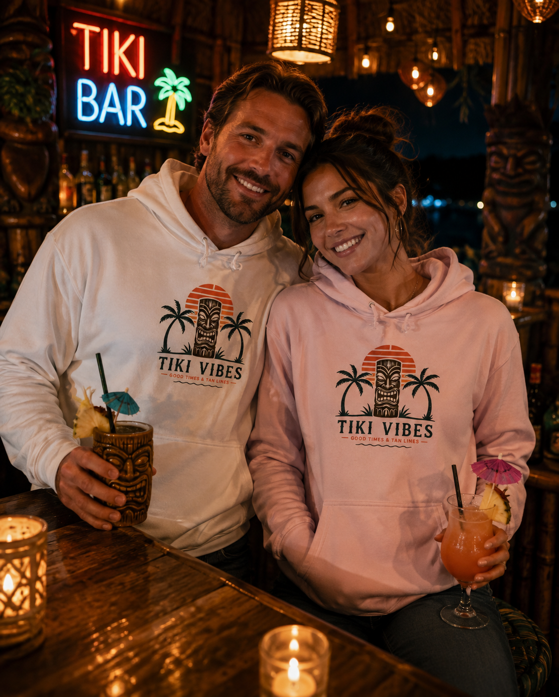 A man and a woman sitting together at a bar, both wearing matching light-colored hoodies with a tropical tiki graphic that reads 'TIKI VIBES'. The man wears a white hoodie and holds a tiki mug, while the woman wears a light pink hoodie and holds a cocktail glass.
