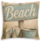 A square decorative throw pillow featuring a rustic beach-themed print with a weathered blue sign that reads 'Beach,' sand, grass, flip-flops, and a metal bucket on a cream background.