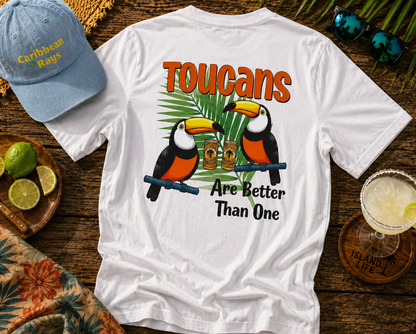 White t-shirt with toucan design and text on a wooden surface with a cap and drink.