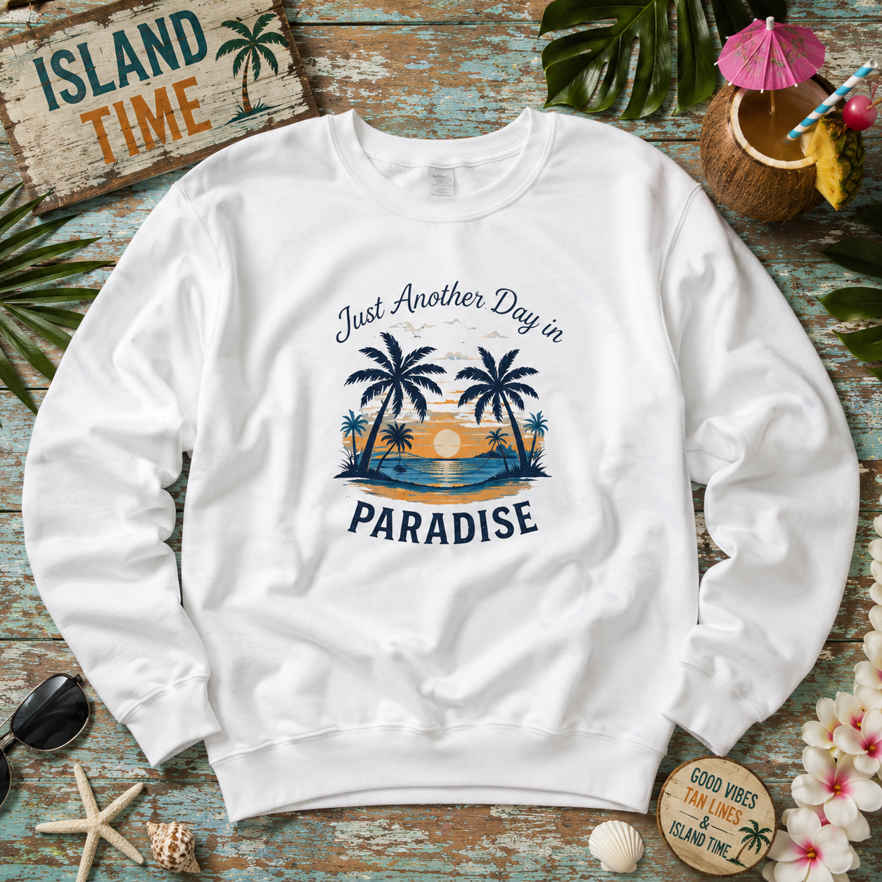 A white crewneck sweatshirt with a tropical beach graphic and the text 'Just Another Day in PARADISE', laid flat on a rustic wooden surface surrounded by beach-themed props like sunglasses, a coconut drink, starfish, shells, and a sign reading 'ISLAND TIME'.