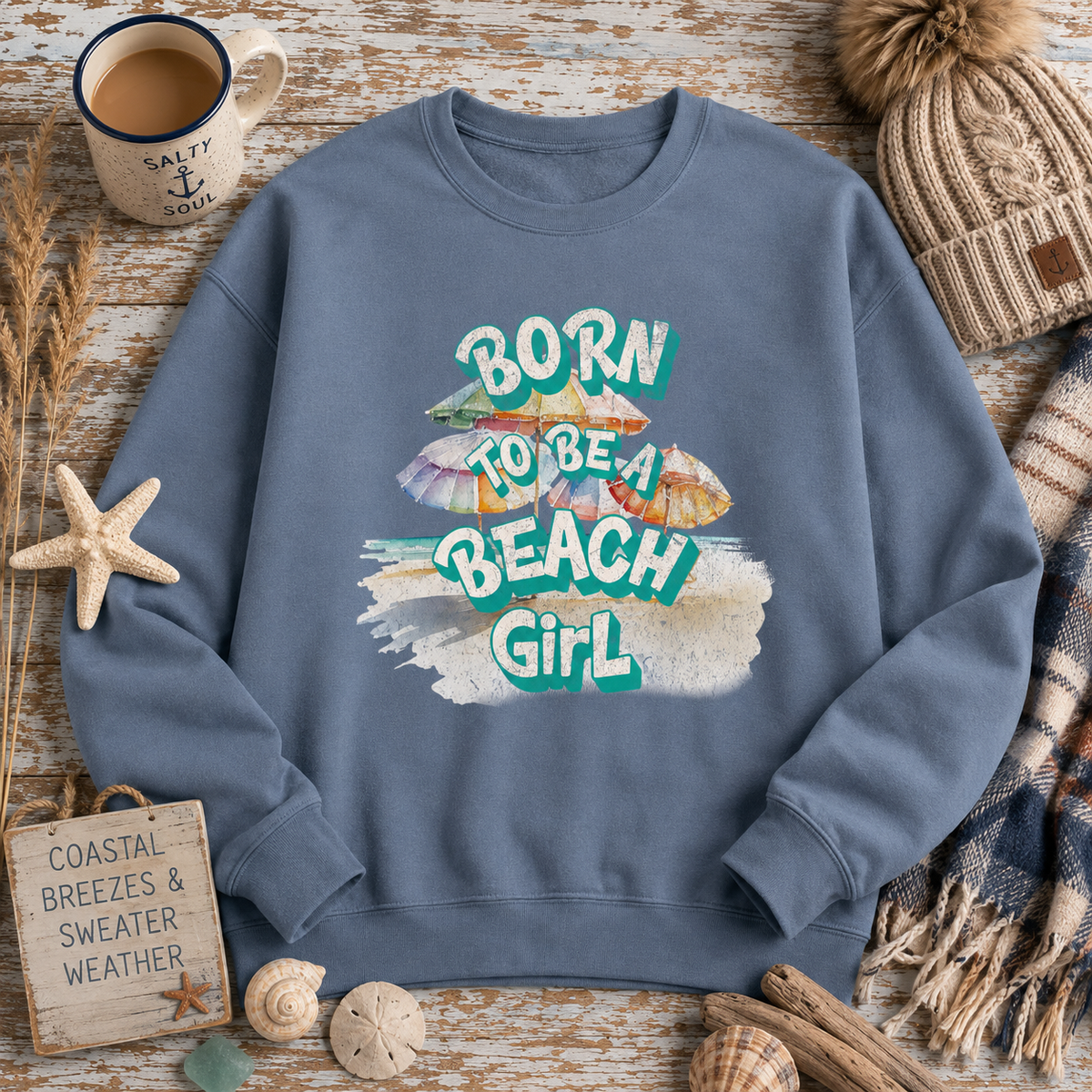 A slate blue crewneck sweatshirt with a colorful graphic reading 'BORN TO BE A BEACH GIRL' surrounded by beach-themed props including a starfish, shells, a mug, and a knit hat on a rustic wood background.