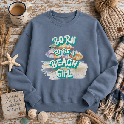 A slate blue crewneck sweatshirt with a colorful graphic reading 'BORN TO BE A BEACH GIRL' surrounded by beach-themed props including a starfish, shells, a mug, and a knit hat on a rustic wood background.