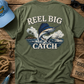 Green t-shirt with 'Reel Big Catch' design on a wooden surface with fishing-themed accessories.