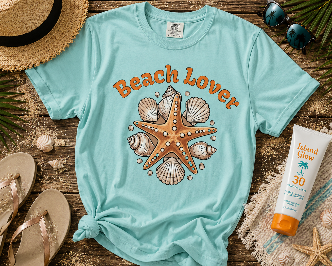 Light blue t-shirt with 'Beach Lover' design featuring a starfish and shells, surrounded by beach accessories on a wooden surface.