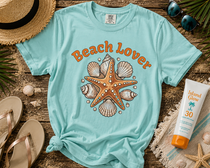 Light blue t-shirt with 'Beach Lover' design featuring a starfish and shells, surrounded by beach accessories on a wooden surface.