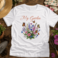 White t-shirt with floral design and 'My Garden' text on a wooden surface with gardening tools and plants.