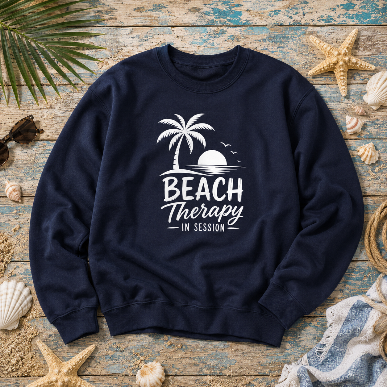 A navy blue crewneck sweatshirt with a white graphic reading 'BEACH Therapy IN SESSION' beneath a palm tree and sunset, laid flat on a rustic blue wooden surface surrounded by seashells, starfish, sunglasses, and a striped towel.