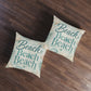 Square floor pillow set with teal “Beach Beach” print and seashell accents, two on wood floor