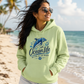 A woman wearing a light green hoodie with a blue dolphin graphic and the text 'Ocean Life Better At The Beach' stands on a sunny beach with palm trees and ocean waves in the background.