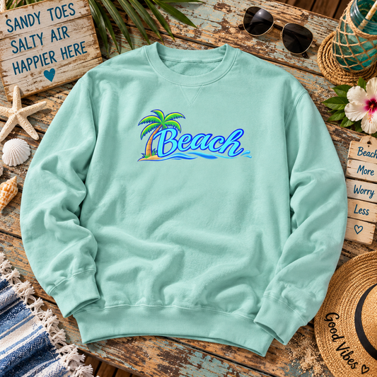 A mint green crewneck sweatshirt with a colorful 'Beach' palm tree graphic on the chest, styled in a beach-themed flat lay on a rustic wooden table with sunglasses, a straw hat, and seashells.