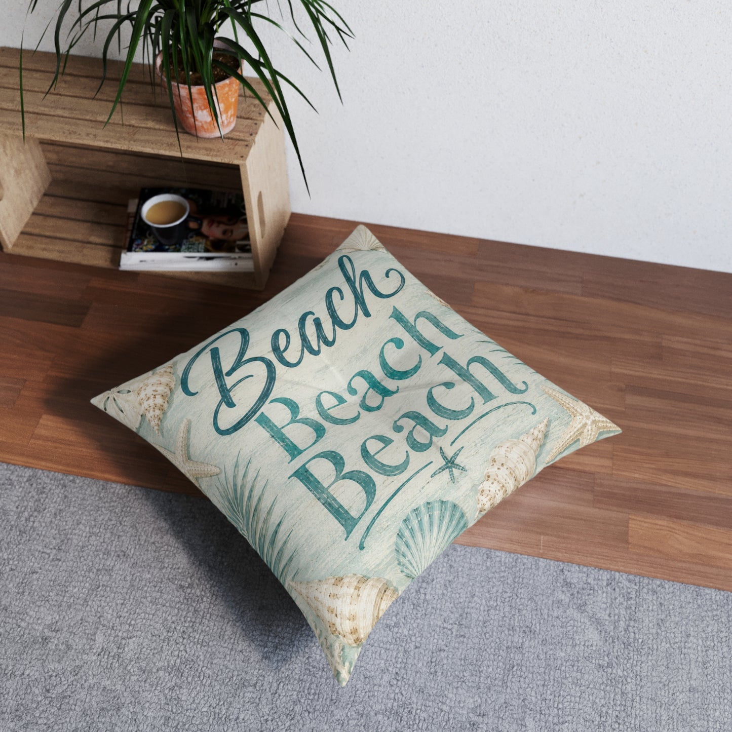 Square floor pillow with teal “Beach” text and seashell pattern, angled on living room floor