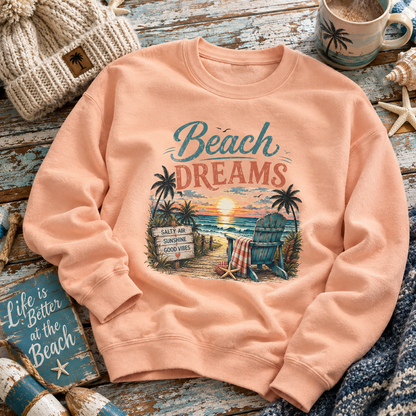 A peach-colored crewneck sweatshirt with a retro beach graphic that reads 'Beach DREAMS' above a sunset beach scene with palm trees, a chair, and a sign. The sweatshirt is styled in a flat-lay on a rustic blue wooden surface with a knit hat, mug, and starfish.