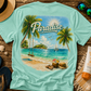 Teal t-shirt with a tropical paradise graphic on a wooden surface with beach accessories.