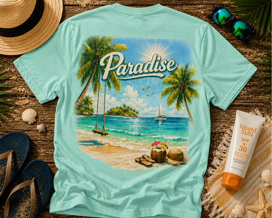 Teal t-shirt with a tropical paradise graphic on a wooden surface with beach accessories.