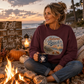A woman sits cross-legged by a campfire at the beach, wearing a maroon sweatshirt with a circular 'Autumn Beach Life' graphic. She holds a navy blue mug, with pumpkins, a lantern, and a wooden sign reading 'Fall is better by the Beach' in the background.
