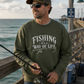 A man wearing an olive green crewneck sweatshirt with the text 'FISHING it's a WAY OF LIFE' stands on a wooden pier holding a fishing rod, with the ocean and sky in the background.