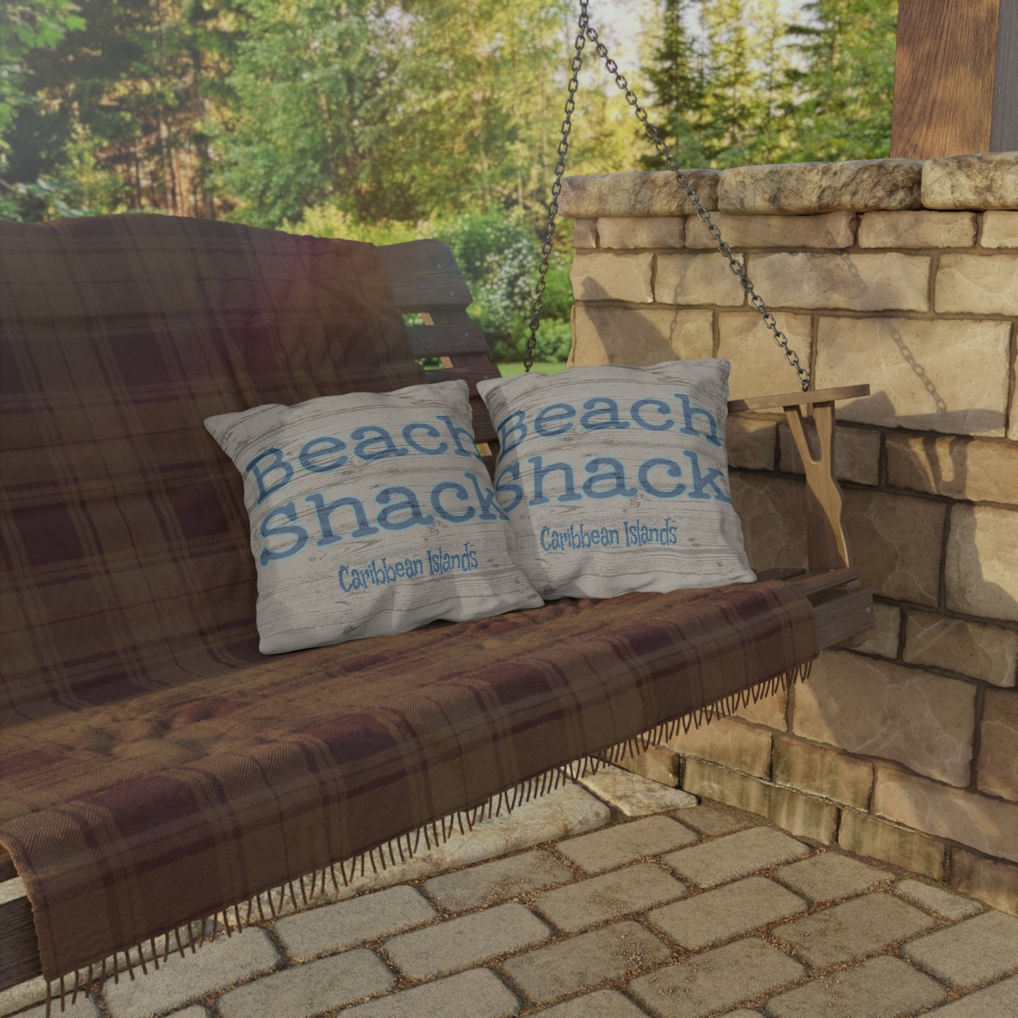 Wooden bench with plaid blanket and 'Beach Shack' pillows against a stone wall and greenery.
