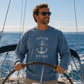 A man wearing a blue crewneck sweatshirt with a white anchor graphic and the text 'FOLLOW THE STARS' stands behind a wooden wheel on a sailboat, with the ocean and blue sky in the background.