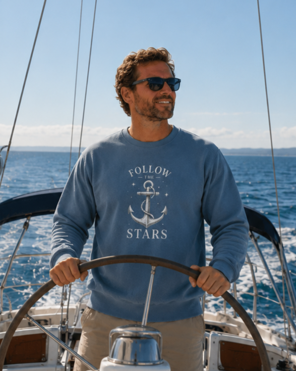 A man wearing a blue crewneck sweatshirt with a white anchor graphic and the text 'FOLLOW THE STARS' stands behind a wooden wheel on a sailboat, with the ocean and blue sky in the background.