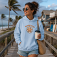A woman stands on a wooden boardwalk at the beach wearing a light blue hoodie with a starfish graphic and the words 'Beach Lover' printed on the front. She is also wearing denim shorts and carrying a woven straw tote bag while holding a coffee cup.