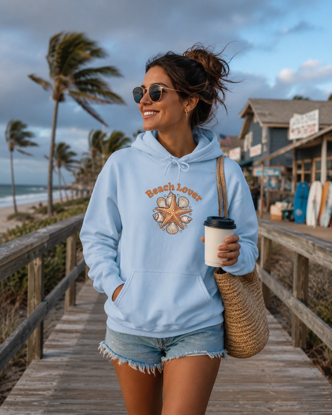 A woman stands on a wooden boardwalk at the beach wearing a light blue hoodie with a starfish graphic and the words 'Beach Lover' printed on the front. She is also wearing denim shorts and carrying a woven straw tote bag while holding a coffee cup.