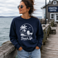 A woman wearing a navy blue crewneck sweatshirt with a white 'Beach Life' palm tree graphic, standing on a wooden pier next to the ocean with a 'COASTAL COFFEE' shop in the background.