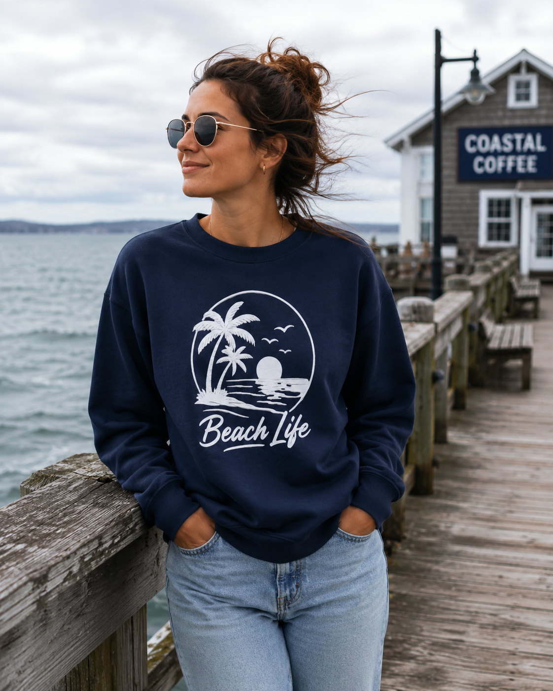 A woman wearing a navy blue crewneck sweatshirt with a white 'Beach Life' palm tree graphic, standing on a wooden pier next to the ocean with a 'COASTAL COFFEE' shop in the background.