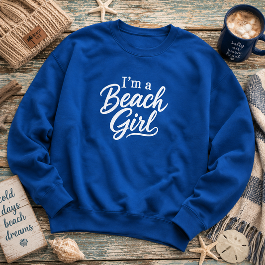 A royal blue crewneck sweatshirt with 'I'm a Beach Girl' printed in white script across the chest, styled in a flat lay on a rustic wooden surface with beach-themed props including a mug, starfish, sand, and woven textiles.