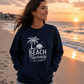 A woman stands on a beach at sunset wearing a navy blue sweatshirt with a white graphic that reads 'BEACH Therapy IN SESSION' with a palm tree and sun design, paired with light denim shorts.